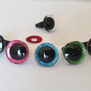 Puede incluir: Un conjunto de ojos de seguridad para manualidades, cada uno con una pupila negra grande rodeada por un iris brillante con purpurina en colores como plata, rosa, azul, verde y oro. Cada ojo tiene un poste de pl&aacute;stico negro y una arandela.
