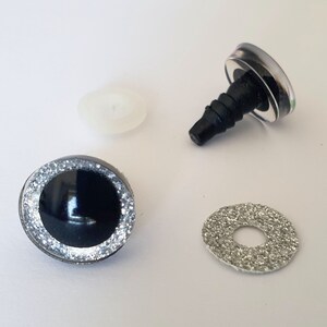 Puede incluir: Un conjunto de ojos para manualidades con un centro negro brillante rodeado por un anillo de purpurina plateada. Los componentes incluyen un ojo de seguridad de pl&aacute;stico con un v&aacute;stago roscado, una arandela de pl&aacute;stico blanco transl&uacute;cido y una arandela de purpurina plateada.