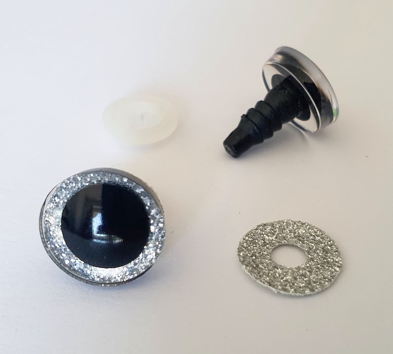 Puede incluir: Un conjunto de ojos para manualidades con un centro negro brillante rodeado por un anillo de purpurina plateada. Los componentes incluyen un ojo de seguridad de pl&aacute;stico con un v&aacute;stago roscado, una arandela de pl&aacute;stico blanco transl&uacute;cido y una arandela de purpurina plateada.