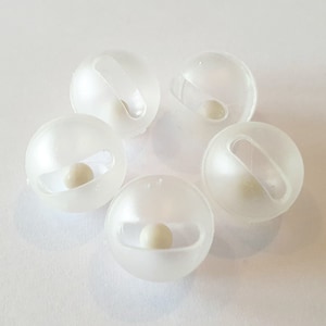 May include: Five translucent, spherical objects, each containing a small, off-white sphere. Each sphere has a rectangular opening and a small handle-like protrusion. The objects are arranged on a white surface.