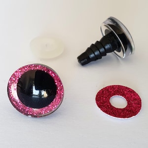 Puede incluir: Dos ojos de seguridad redondos con pupilas negras y bordes de iris con purpurina rosa, con tallos de pl&aacute;stico negro para su fijaci&oacute;n, acompa&ntilde;ados de una arandela de pl&aacute;stico transl&uacute;cido y un inserto circular de purpurina roja.