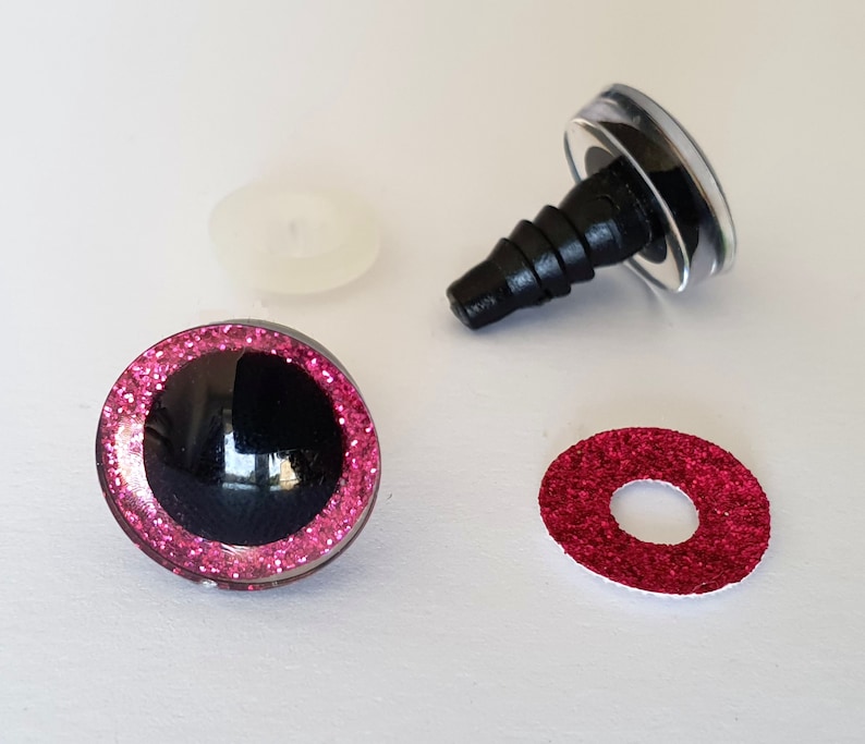 Puede incluir: Dos ojos de seguridad redondos con pupilas negras y bordes de iris con purpurina rosa, con tallos de pl&aacute;stico negro para su fijaci&oacute;n, acompa&ntilde;ados de una arandela de pl&aacute;stico transl&uacute;cido y un inserto circular de purpurina roja.