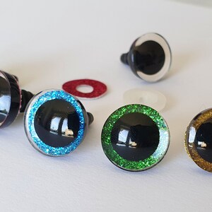 Puede incluir: Un conjunto de ojos de seguridad redondos para manualidades, con pupilas negras rodeadas de iris brillantes en rosa, azul, verde y dorado, junto con un ojo transparente y dos peque&ntilde;as arandelas circulares de pl&aacute;stico rojo y blanco transl&uacute;cido.
