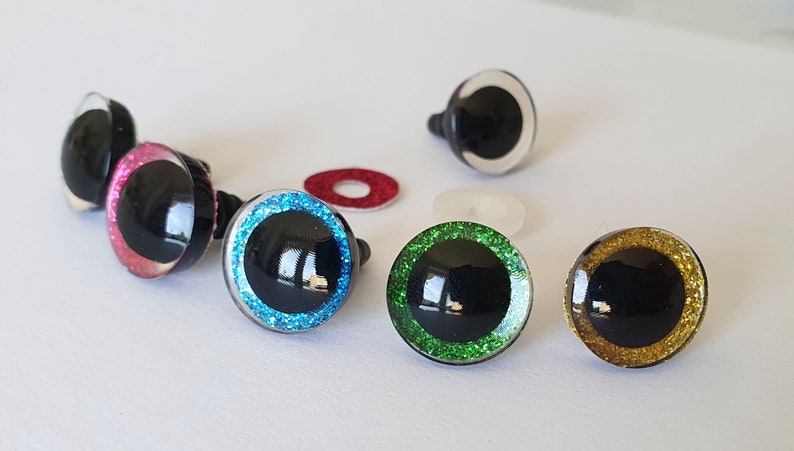 Puede incluir: Un conjunto de ojos de seguridad redondos para manualidades, con pupilas negras rodeadas de iris brillantes en rosa, azul, verde y dorado, junto con un ojo transparente y dos peque&ntilde;as arandelas circulares de pl&aacute;stico rojo y blanco transl&uacute;cido.