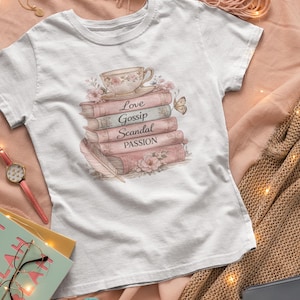 May include: White t-shirt featuring a stack of books with the words "Love, Gossip, Scandal, Passion" and a teacup illustration. The design includes floral and butterfly accents. The shirt is displayed on a pink blanket.