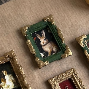 May include: Small framed artworks with gold detailing. One frame displays a rabbit image, another a child's portrait, and a third a floral pattern. The frames have green or red backgrounds and are set on a neutral surface.