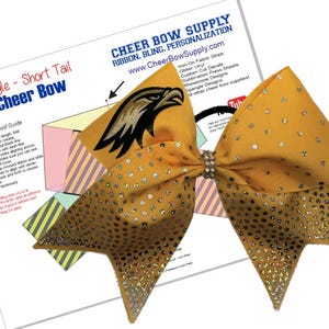 May include: A gold cheer bow with a short tail and rhinestones. The bow is decorated with a black and gold eagle graphic. The image also includes a fold guide with instructions on how to make a cheer bow.