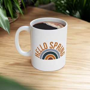 May include: White ceramic mug filled with dark liquid, featuring the text "HELLO SPRING" in brown and a rainbow design. The mug sits on a wooden surface.