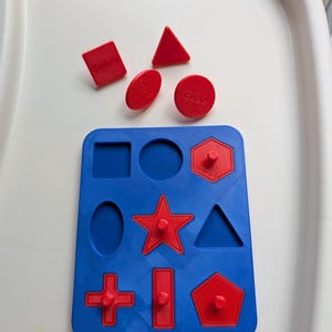 May include: A blue shape sorter toy with red shapes. The shapes include a square, oval, triangle, circle, star, rectangle, cross, and pentagon. The shapes have pegs to fit into the corresponding holes.