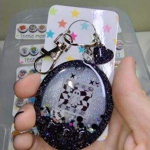 May include: A black glitter resin keychain with a clear center, featuring a ghost design on a checkered background. The keychain includes a silver clasp, a heart charm, and small decorative elements. The text "RIP" is visible.