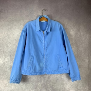 May include: A light blue jacket with a collared neckline and a full-length zipper. The jacket has two front pockets and elastic cuffs and hem. The jacket is hanging on a wooden hanger.