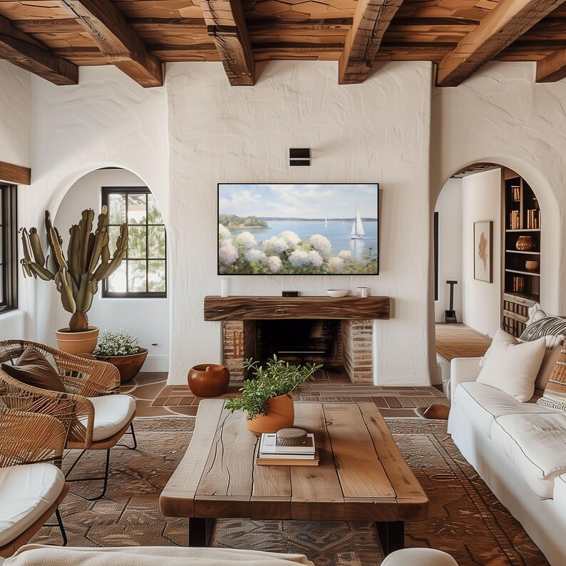 May include: A cozy living room with a fireplace, a large wooden coffee table, and two wicker chairs. A TV displays a scenic view. The room features a neutral color palette with wooden beams and a cactus plant.