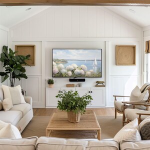 May include: A bright, airy living room with white walls, a large flat-screen TV, and a woven coffee table. The room features a neutral color palette with a large sofa, armchairs, and decorative pillows. A painting of a seascape hangs above the white cabinet.