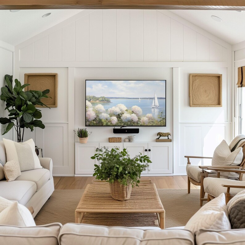 May include: A bright, airy living room with white walls, a large flat-screen TV, and a woven coffee table. The room features a neutral color palette with a large sofa, armchairs, and decorative pillows. A painting of a seascape hangs above the white cabinet.