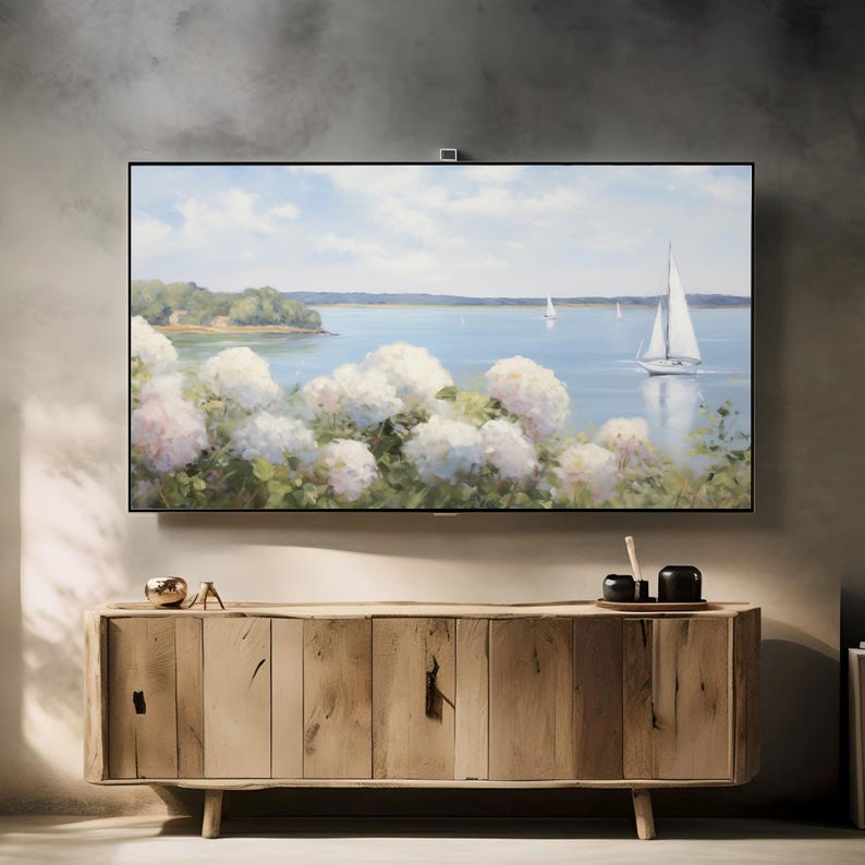 May include: A large, flat-screen television displays a serene landscape painting of a sailboat on the water, with white flowers in the foreground. The TV is mounted above a light-colored wooden cabinet with a natural finish. Decorative items sit on top of the cabinet.