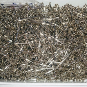 May include: A close-up shot of a box overflowing with shiny, silver-colored pins. The pins are densely packed, with their sharp points and rounded heads creating a textured surface. The pins appear to be new and unused.