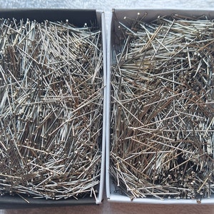 May include: Two rectangular boxes filled with silver sewing pins. The pins are scattered and appear to be made of metal. One box is black, and the other is white. The pins have small, round heads.