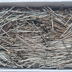 May include: A box filled with numerous silver sewing pins. Each pin has a small, round head and a sharp, pointed tip. The pins are scattered throughout the box, creating a dense, metallic texture. The box is white.