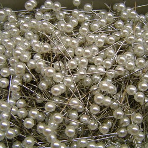 May include: A collection of pearl-headed pins with silver shafts. The pins are densely packed together, creating a textured surface. The pearls are a creamy white color, and the pins are likely used for sewing or crafting projects.