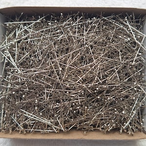 May include: A box filled with numerous silver sewing pins. The pins are scattered and appear to be made of metal, with small, round heads. The box is white with a cardboard base, suggesting a craft or sewing supply.