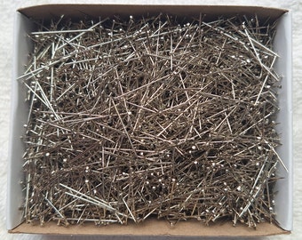 May include: A box filled with numerous silver sewing pins. The pins are scattered and appear to be made of metal, with small, round heads. The box is white with a cardboard base, suggesting a craft or sewing supply.