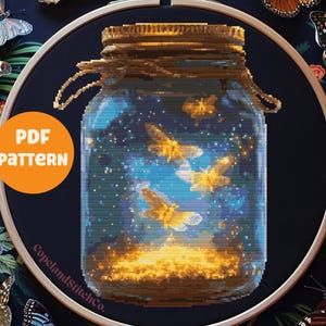 Può includere: Un barattolo di vetro con un coperchio, pieno di lucciole luminose e un cielo notturno stellato. Il barattolo è circondato da un telaio da ricamo in legno e foglie verdi.