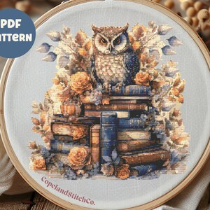 May include: A cross stitch pattern featuring an owl perched on a stack of books with autumnal foliage and flowers. The pattern is in a round hoop and includes the text "CopelandStitchCo."
