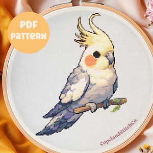 以下が含まれることがあります： 枝に止まっているコザクラインコのクロスステッチパターン。コザクラインコは灰色と白で、黄色い冠羽と桃色の頬の斑点があります。パターンは、白の刺繍枠の中にあり、桃色の円に「PDF PATTERN」というテキストと、下に「CopelandStitchCo.」というテキストがあります。