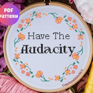 May include: A cross-stitch embroidery hoop with the words "Have The Audacity" in black. The design is surrounded by a floral border in orange and green. The words "PDF PATTERN" are in a pink and purple circle.