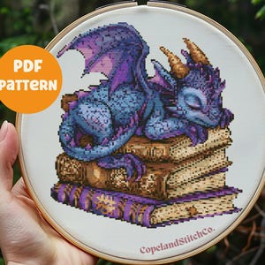 May include: A purple and blue dragon with closed eyes is sleeping on a stack of four books. The dragon has a long tail and large wings. The books are brown with purple covers. The image is a cross stitch pattern.