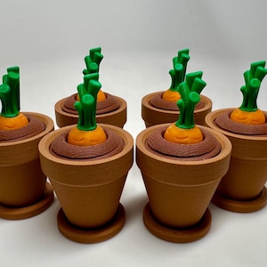 Puede incluir: Seis pequeñas macetas de terracota con tapas verdes y caras de zanahoria naranja. Las macetas están dispuestas en semicírculo sobre una superficie blanca.