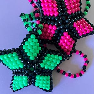 Könnte beinhalten: Zwei sternförmige Perlenornamente. Eines ist schwarz und grün, das andere ist schwarz und rosa. Die Perlen sind so angeordnet, dass sie ein Sternmuster bilden. Jedes Ornament hat eine Schlaufe aus Perlen.