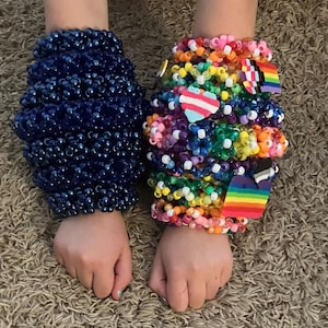 Könnte beinhalten: Zwei Armbänder aus Perlen. Eines ist tiefblau, das andere ist ein Regenbogen aus Farben. Das Regenbogenarmband hat ein Herz und eine Regenbogenflagge. Die Armbänder bestehen aus kleinen Perlen.