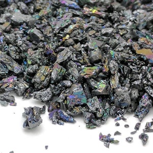 May include: A close-up of a pile of iridescent silicon carbide crystals. The crystals are dark gray with flashes of rainbow colors, including blue, green, and purple. The crystals have a rough, fractured appearance.