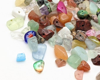 May include: A collection of assorted gemstone beads in various colors, including clear, green, orange, red, and blue. The beads are tumbled and have holes for stringing. Some beads are opaque, while others are translucent.