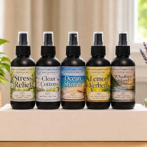 May include: Five black spray bottles of room and linen spray are displayed on a white surface. Each bottle has a different label: Stress Relief, Clean Cotton, Ocean Shores, Lemon Verbena, and Whidbey Island. The bottles are 4 oz each.