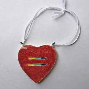 May include: A red heart-shaped pendant with a white ribbon. The pendant features two horizontal lines in yellow, green, and blue. The pendant is likely a symbol of equality and is suitable for wearing as a necklace or ornament.