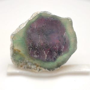 May include: A raw, natural fluorite gemstone with a vibrant color gradient. The center is a deep purple, surrounded by a ring of green, and edged with a lighter green hue. The stone has a rough, textured surface.