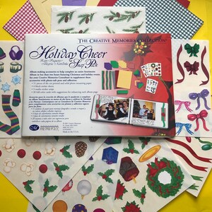 May include: A red, white, and green holiday scrapbook kit with the title "Holiday Cheer Snap Pack" and a photo of a family in front of a Christmas tree. The kit includes stickers, paper, and other embellishments.