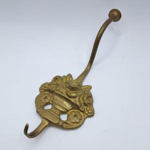 Op de afbeelding: Een messing wandhaak met een decoratieve basisplaat met een reliëfontwerp van een vaas gevuld met bloemen. De haak heeft een lange, gebogen bovenarm die eindigt in een kleine ronde knop en een kortere, kleinere onderste haak aan de basis.