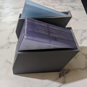 Può includere: Due scatole portaoggetti grigie angolate, una impilata sull'altra. La scatola superiore contiene un elemento triangolare azzurro. La scatola inferiore è riempita con una pila di custodie per carte di plastica trasparente. Le scatole sono su una superficie di marmo bianco.