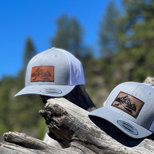 May include: Two gray and white baseball caps with a brown leather patch featuring a mountain scene. The caps are sitting on a weathered wooden log.