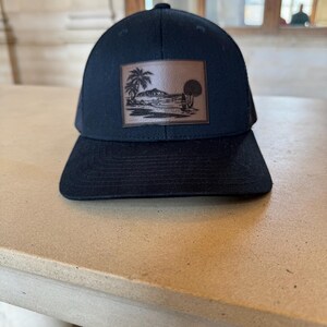 May include: A black trucker hat with a brown leather patch featuring a palm tree and island scene.
