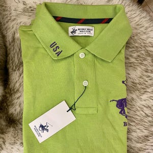 May include: A green polo shirt with a purple polo player logo and the text "Beverly Hills Polo Club" on the front. The shirt also has a red and black striped collar and the text "USA" embroidered on the left side of the collar.