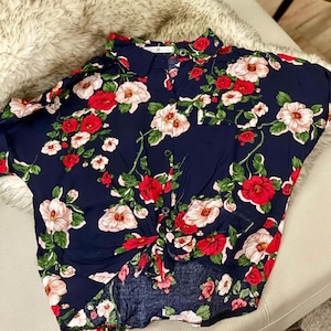 May include: Navy blue button-down shirt with a vibrant floral pattern. The shirt features a mix of red, white, and pink flowers with green leaves. The shirt is knotted at the waist and has short sleeves.