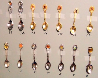 May include: A collection of 18 silver and gold souvenir spoons with various designs. The spoons are arranged in two rows on a light grey surface. Each spoon has a different design, including a person, a building, and a flag. The spoons are numbered 1 through 18.