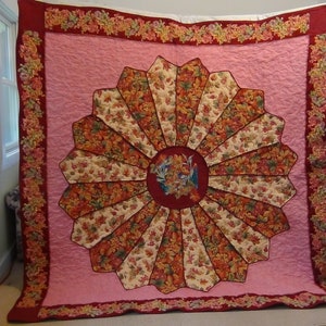 May include: A pink and burgundy quilt with a floral pattern. The center of the quilt features a large, circular design with a burgundy center and a radiating pattern of beige, brown, and red.