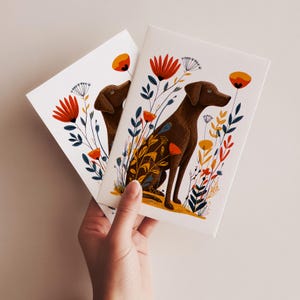 May include: Two white rectangular greeting cards featuring a brown dog illustration surrounded by stylized red, orange, and dark blue flowers and foliage. The dog is depicted in a seated position, facing right, amidst a colorful botanical arrangement.