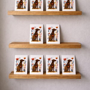 May include: Ten identical rectangular greeting cards featuring a brown dog sitting in profile, surrounded by stylized red, orange, and blue floral and botanical illustrations, arranged on three horizontal wooden shelves against a plain wall.