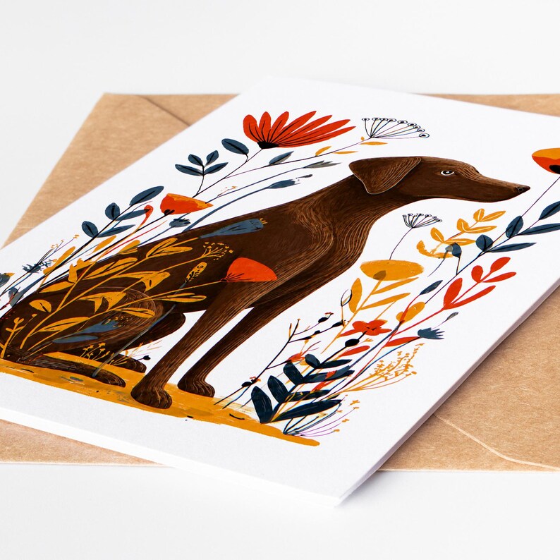 May include: A rectangular greeting card featuring a brown dog illustration surrounded by stylized orange, red, yellow, and dark blue flowers and foliage. The card is positioned on top of a light brown paper envelope on a white surface.